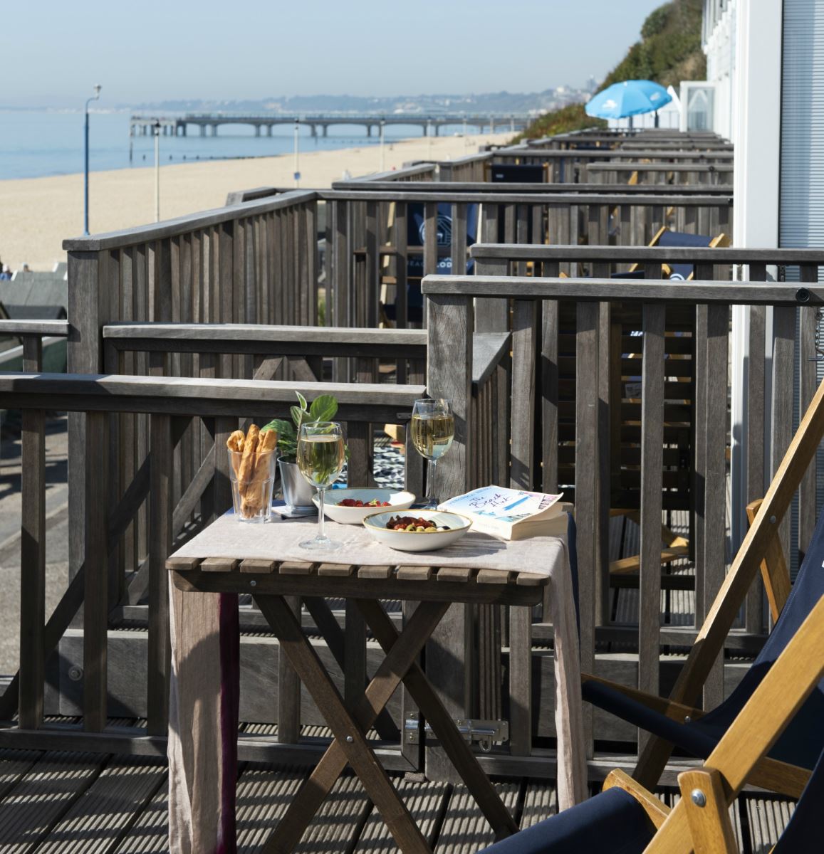 An outside shot of the Beach Lodges on a bright and warm day, featuring two deck chairs with a table featuring small dishes of olives and strawberries, bread sticks and two glasses of wine