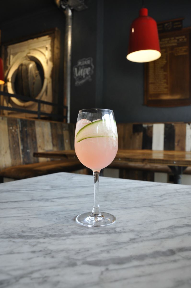 A single cocktail stood on a table in The Dancing Moose, Bournemouth. The drink is pink in colour with a green hollow leaf immersed in it. 