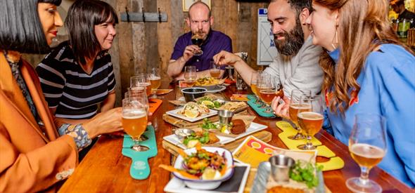 Five people around a table enjoying a selection of food and beer.