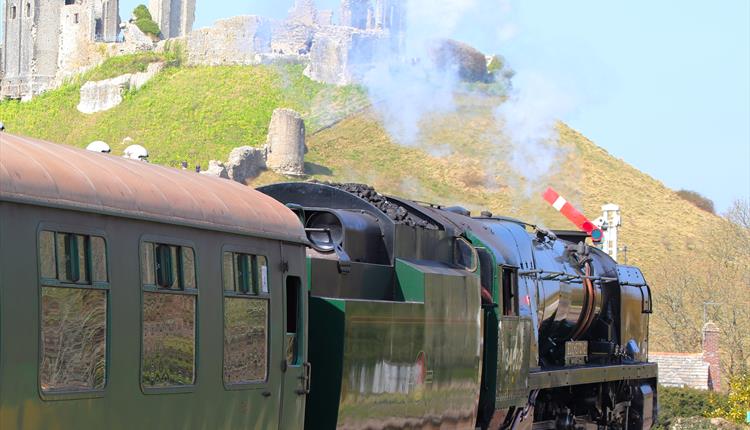 a green train from a side angle with a caste ruins on a hill overlooking the train