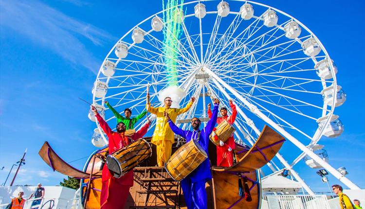 5 men playing instruments whilst dressed in colourful clothing in front of Bournemouths big wheel