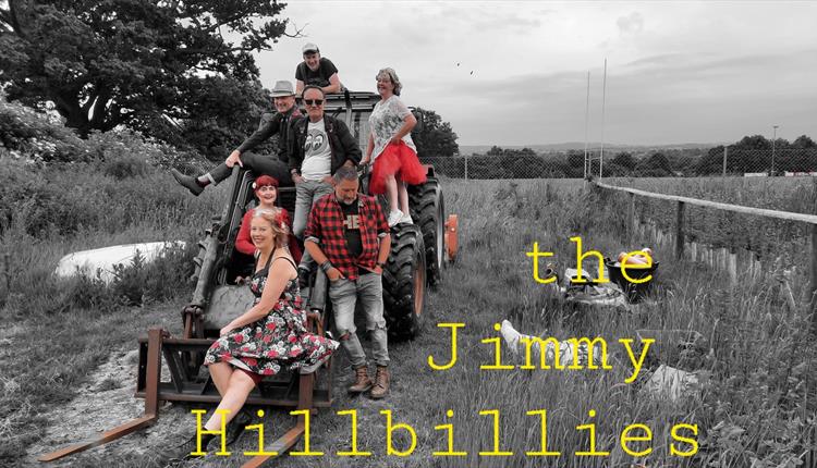 Four men and three women posed on a digger in a field. Background is black and white but the figures are in colour