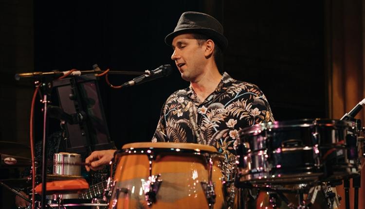 Young man playing drums and singing into a mic wearing a hat and colourful shirt.